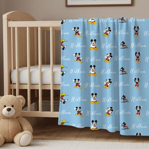 Puede incluir: Una manta de beb&eacute; azul claro con un patr&oacute;n de Mickey Mouse en varias poses. El nombre "William" est&aacute; escrito en cursiva en la manta. Un oso de peluche est&aacute; frente a una cuna de madera.