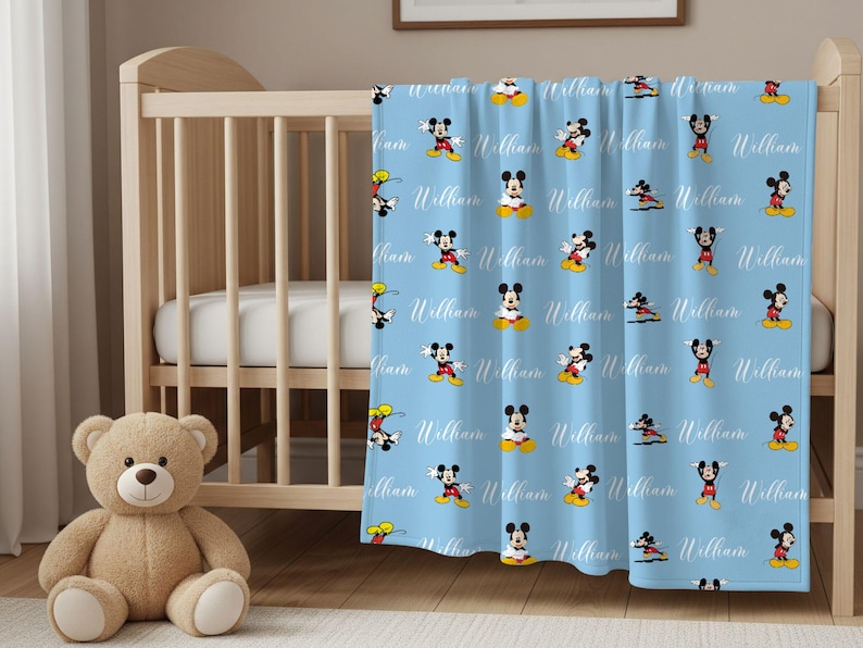Puede incluir: Una manta de beb&eacute; azul claro con un patr&oacute;n de Mickey Mouse en varias poses. El nombre "William" est&aacute; escrito en cursiva en la manta. Un oso de peluche est&aacute; frente a una cuna de madera.