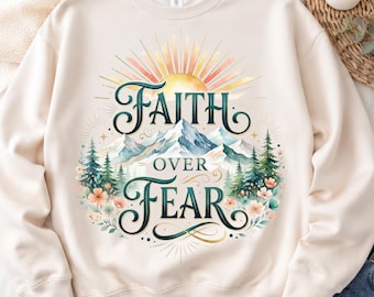 Pode incluir: Uma camisola de gola redonda cor de creme com uma impressão gráfica de um sol a nascer sobre montanhas cobertas de neve e pinheiros. O texto "FAITH OVER FEAR" está impresso em letras estilizadas verde-escuras, rodeado por pequenas flores.