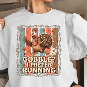 May include: A white crewneck sweatshirt featuring a graphic of a brown turkey running with a surprised expression. The design includes vertical stripes, autumn leaves, acorns, and the text GOBBLE? I PREFER RUNNING in a decorative frame.