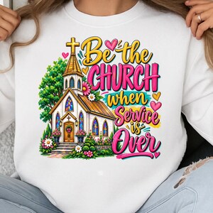 Pode incluir: Uma camisola branca com um gr&aacute;fico colorido de uma igreja com torre, rodeada por &aacute;rvores verdes e flores cor-de-rosa e brancas. O texto diz, Be the Church when Service is Over, em letras estilizadas amarelas e cor-de-rosa.