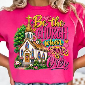 Pode incluir: Uma camisola cor-de-rosa brilhante com uma ilustra&ccedil;&atilde;o gr&aacute;fica de uma igreja com vitrais, rodeada por &aacute;rvores verdes e flores coloridas. O texto diz Be the CHURCH when Service is Over em letras amarelas e cor-de-rosa.
