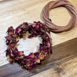 May include: Two circular rings made of twisted natural vine material. One ring is bare, showing a light brown woody texture, while the other ring is densely covered with dried, deep magenta and dark purple rose petals attached to the vine base.