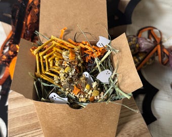 Könnte beinhalten: Eine quadratische braune Pappschachtel gefüllt mit getrocknetem Heu, orangen Papierschnipseln und kleinen Kräuterleckerlis. Die Schachtel enthält ein gewebtes Spinnennetz aus Zweigen, getrocknete Kamillenblüten und kleine weiße Papiergespenster.