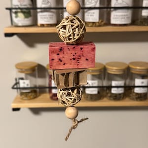 May include: A hanging bird toy featuring a natural wood and woven ball design. The toy includes a red-stained wooden block and a cardboard piece. The toy is suspended by a natural twine string, with wooden beads. The background shows spice jars.