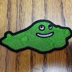 Może przedstawiać: Zielona haftowana łatka przedstawiająca uśmiechniętą, kreskówkową plamę z czarnymi konturami i białymi oczami.