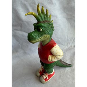 May include: A green dinosaur figurine wearing a red and white varsity jacket and red sneakers. The dinosaur has a green crest and a long tail. The figurine is standing on a white surface.