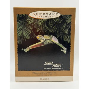 Könnte beinhalten: Eine rechteckige Schachtel für ein Keepsake-Ornament mit einem Klingon Bird of Prey Raumschiff aus Star Trek The Next Generation. Die Schachtel zeigt das Schiff vor einem Tannenzweighintergrund mit dem Text Star Trek The Next Generation und Magic.