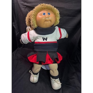 Può includere: Una bambola vintage con capelli biondi di lana, occhi blu e carnagione abbronzata. La bambola indossa un completo da cheerleader bianco e nero con una gonna rossa e la lettera "W" sul petto. Le scarpe da ginnastica bianche completano il look.