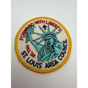 Puede incluir: Un parche bordado circular con borde amarillo sobre fondo blanco. Presenta la Estatua de la Libertad en color turquesa, una pequeña flor de lis azul oscuro y texto en rojo y azul oscuro: FORWARD WITH LIBERTY, FALL '86 y ST LOUIS AREA COUNCIL.