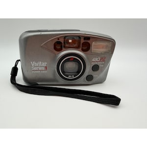 May include: A silver Vivitar Series 1 480PZ Power Zoom 35mm film camera with a black wrist strap. The front features a 38-70mm lens, a built-in flash, and text reading Vivitar Series 1 Power Zoom and 480PZ Data Back with Tele and Wide buttons.