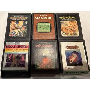 May include: Six vintage Atari game cartridges are displayed, including "Missile Command," "Stampede," "Video Olympics," "Riddle of the Sphinx," "Star Raiders," and "Journey Escape." The cartridges have colourful graphics and text, representing classic video game art.