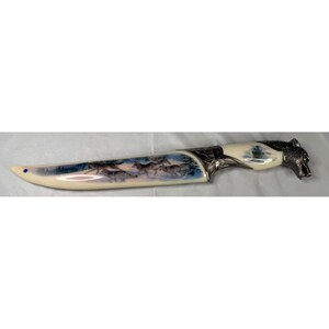 Puede incluir: Un cuchillo decorativo con mango y hoja color crema. La hoja presenta una escena pintada de lobos en un paisaje nevado. El mango está adornado con una cabeza de lobo plateada y detalles metálicos decorativos.