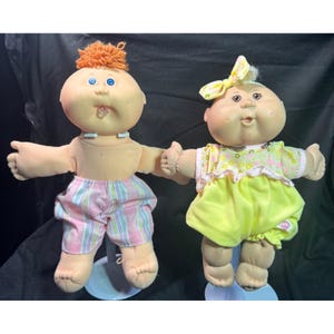 Könnte beinhalten: Zwei Vintage Cabbage Patch Kids Puppen. Eine Puppe hat orangefarbenes Haar, blaue Augen und gestreifte rosa und weiße Shorts. Die andere Puppe hat blondes Haar, eine gelbe Schleife und ein gelbes Kleid mit Blumenmuster. Beide Puppen halten sich an den Händen.