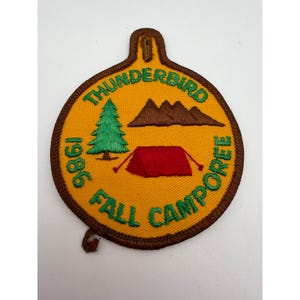May include: A circular embroidered patch with a yellow background and brown border. It features a green pine tree, brown mountains, and a red tent. The text reads THUNDERBIRD 1986 FALL CAMPOREE in green letters. The patch has a small tab at the top.