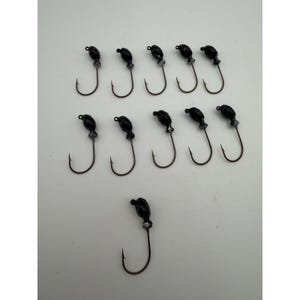Puede incluir: Once cabezas de anzuelo para pesca dispuestas en dos filas de cinco con una centrada debajo. Cada cabeza de anzuelo presenta una parte superior redondeada de color negro brillante unida a un anzuelo metálico curvo con un cuello plateado.