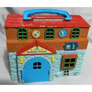 May include: A colorful toy playset featuring a building with a red roof, brown wooden facade, and blue stone base. The playset has a blue handle, arched windows, and the text "SODOR ENGINE WORKS" and "ENGINE WASH".