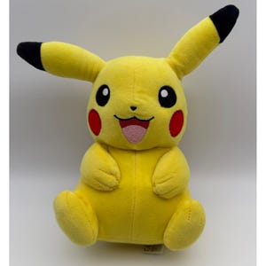 May include: A yellow plush toy featuring the character Pikachu with long ears tipped in black, round red cheeks, and a smiling face with an open mouth showing a pink tongue. The soft stuffed figure has black eyes with white highlights and small paws.