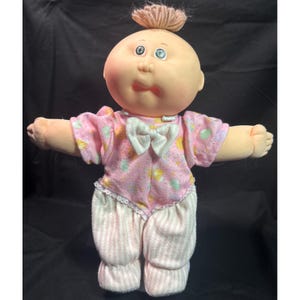 Op de afbeelding: Een vintage Cabbage Patch Kid-pop met een perzikkleurig gezicht, blauwe ogen en een kleine pluk lichtbruin garen haar. De pop draagt een roze onesie met een witte strik en een patroon van gele en groene vormen, gecombineerd met een gestreepte broek.