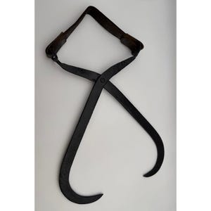 May include: A pair of vintage black iron ice tongs featuring two curved, pointed gripping arms connected by a central pivot point and a sturdy metal handle at the top. The metal surface has a textured, matte finish with visible signs of age.