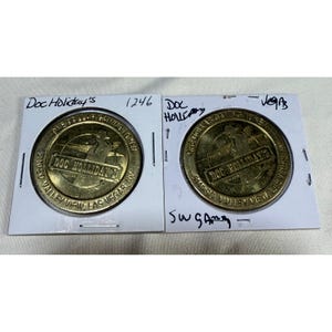 Puede incluir: Dos fichas de juego de un dólar "Doc Holiday's" de color dorado. Las fichas están en fundas protectoras. El texto de las fichas dice "Doc Holiday's" y "One Dollar Gaming Token". Las fichas son de "5010 So. Valley View, Las Vegas, NV".