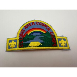 Pode incluir: Um emblema bordado semicircular com fundo azul, um arco-íris sobre uma paisagem verde, uma tenda amarela e um rio. O texto ST. LOUIS AREA COUNCIL CAMPS está bordado em vermelho. Duas flores-de-lis amarelas estão presentes nas laterais.