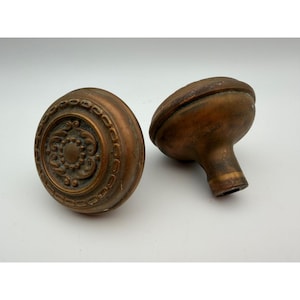 May include: Two round door knobs made of aged bronze metal. The front-facing knob features a central circular medallion surrounded by a decorative border of repeating oval links and a floral relief pattern. Both knobs have a dark, weathered finish.