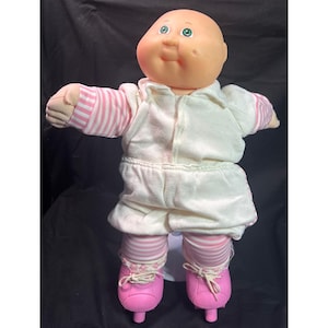 May include: A vintage Cabbage Patch Kid doll wearing a white outfit with pink striped accents and pink roller skates. The doll has a bald head, green eyes, and a smiling expression. The outfit includes a jacket, skirt, and matching striped socks.