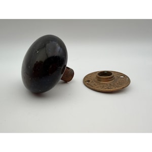 May include: A dark brown, smooth, oval-shaped door knob with a metallic stem, accompanied by a separate circular brass backplate featuring an intricate, embossed floral pattern and two screw holes for mounting on a door surface.
