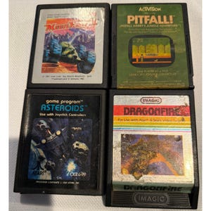 May include: Four vintage Atari video game cartridges. Titles include "Moon Patrol", "Pitfall!", "Asteroids", and "Dragonfire". The cartridges feature colorful graphics and text, with the "Pitfall!" cartridge in a green case.