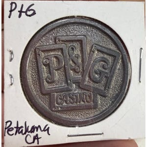 Puede incluir: Ficha de casino plateada con las letras "P & G" dentro de diseños de naipes, con "CASINO" debajo. La ficha está en un tarjetero blanco con texto manuscrito "P+6" y "Petaluma CA".