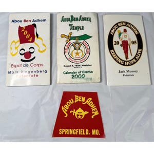 以下が含まれることがあります： Abou Ben Adhemの記念品のコレクション。イベントカレンダーや赤いエンブレムなど。2000年、2004年、2005年などの日付が記載されたテキストとグラフィックが特徴です。アイテムは白と赤です。