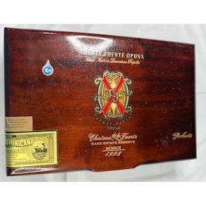 May include: A wooden cigar box with a dark brown finish. The box features gold and red lettering and a decorative emblem. Text on the box includes "Fuente OpusX", "Chateau de la Fuente", and "Rare Estate Reserve".