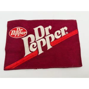 May include: A rectangular maroon fabric patch featuring the white embroidered Dr Pepper logo and a diagonal red stripe. The design includes the classic oval Dr Pepper emblem on the left side with the brand name written in large white script lettering.