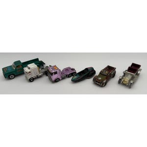 May include: A collection of six die-cast toy vehicles including a green pickup truck, a white semi-truck cab, a metallic pink semi-truck cab, a dark teal and black futuristic vehicle, a weathered brown truck, and a silver vintage-style car.