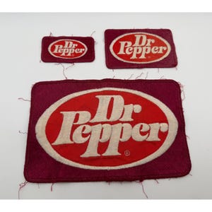 May include: Three rectangular embroidered patches in varying sizes, each featuring a maroon background and a central red oval with the white Dr Pepper logo. The patches have visible loose threads along the edges and a registered trademark symbol.