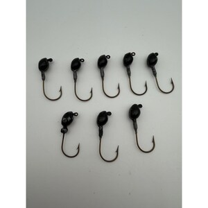 Puede incluir: Un conjunto de ocho cabezas plomadas para pesca, cada una con un peso de plomo negro redondeado unido a un anzuelo de metal curvado. Los anzuelos están dispuestos en dos filas, cinco en la fila superior y tres en la inferior, con un diseño uniforme.