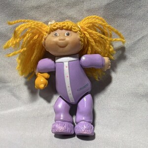 Pode incluir: Uma boneca vintage Cabbage Patch Kids com cabelo amarelo em tranças. A boneca usa um macacão roxo com acabamento branco e uma frente abotoada. Possui luvas amarelas e botas roxas felpudas.