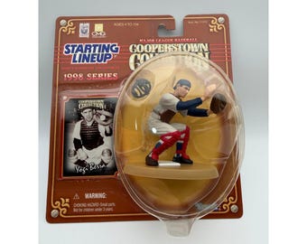 May include: A 1998 Starting Lineup Cooperstown Collection action figure of Yogi Berra in a catcher's crouch, wearing a white uniform with red accents. The figure is encased in a clear plastic bubble on a brown cardboard backing with text.