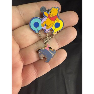 May include: A Winnie the Pooh pin featuring Pooh Bear holding onto three balloons, connected by a chain to an Eeyore pin with a pink bow. The pin is colorful with blue, yellow, and red accents.