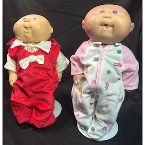 May include: Two vintage Cabbage Patch Kids dolls. One doll wears a red jumpsuit with a white shirt and bow tie. The other doll is in a pink and white onesie with a rainbow and cloud pattern. Both dolls have blue eyes and bald heads.