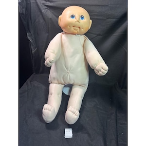 May include: A vintage Cabbage Patch Kid doll with a soft, flesh-toned body and head. The doll has blue eyes, a small nose, and a stitched mouth. Its arms and legs are made of soft fabric. A small tag is visible near the doll's feet.