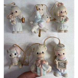 Könnte beinhalten: Eine Sammlung von sechs Precious Moments Porzellanornamenten. Die Ornamente zeigen Figuren von Kindern und Tieren mit goldenen Schnuraufhängern. Jedes Ornament hat eine Jahreszahl aufgedruckt, darunter 1996 und 1997. Die Ornamente sind in verschiedenen Posen.