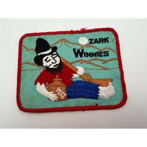 May include: A rectangular embroidered patch with a red border on a light blue background. It features a figure in a black hat, red shirt, and blue pants holding a brown jug labeled "XXX" and a rifle. The text "ZARK WINNIES" appears in black.