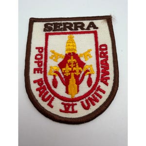 May include: A shield-shaped embroidered patch with a brown border on a cream background. It features the text SERRA POPE PAUL VI UNIT AWARD in red and brown. The center displays a yellow papal tiara and crossed keys in red and yellow thread.