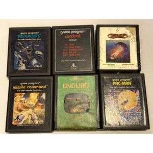 May include: A collection of six vintage Atari game cartridges, including "Asteroids," "Combat," "Journey Escape," "Missile Command," "Enduro," and "Pac-Man." Each cartridge features colorful artwork and game titles.
