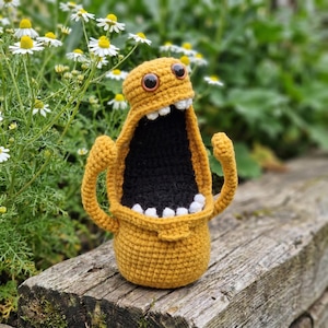 May include: A handmade, mustard yellow crochet monster with a wide open black mouth and white teeth. The monster has two large brown eyes and two arms raised in the air. The monster is sitting on a weathered wooden surface.