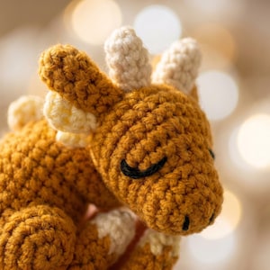 Könnte beinhalten: Handgefertigtes gehäkeltes Giraffen-Spielzeug in Goldbraun- und Cremetönen. Die Giraffe hat geschlossene schwarze Augen, kleine Ohren und cremefarbene Hörner. Das Stofftier ist mit einem strukturierten Stich gefertigt, was ihm ein gemütliches Aussehen verleiht.