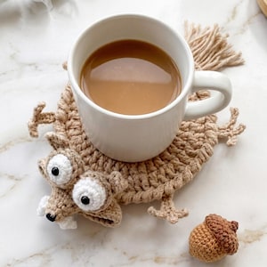 May include: A white mug containing coffee rests on a tan crochet squirrel coaster, featuring large white eyes and a fluffy tail. A small, brown crochet acorn is placed beside the coaster. The coaster is approximately 15 cm in length.