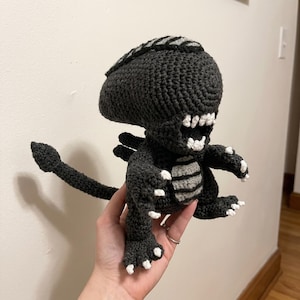 May include: A handmade crochet Xenomorph doll in dark gray with white accents. The creature has a large head, a long tail, and a detailed body. The mouth is open, revealing white teeth. The doll is a unique, handcrafted item.
